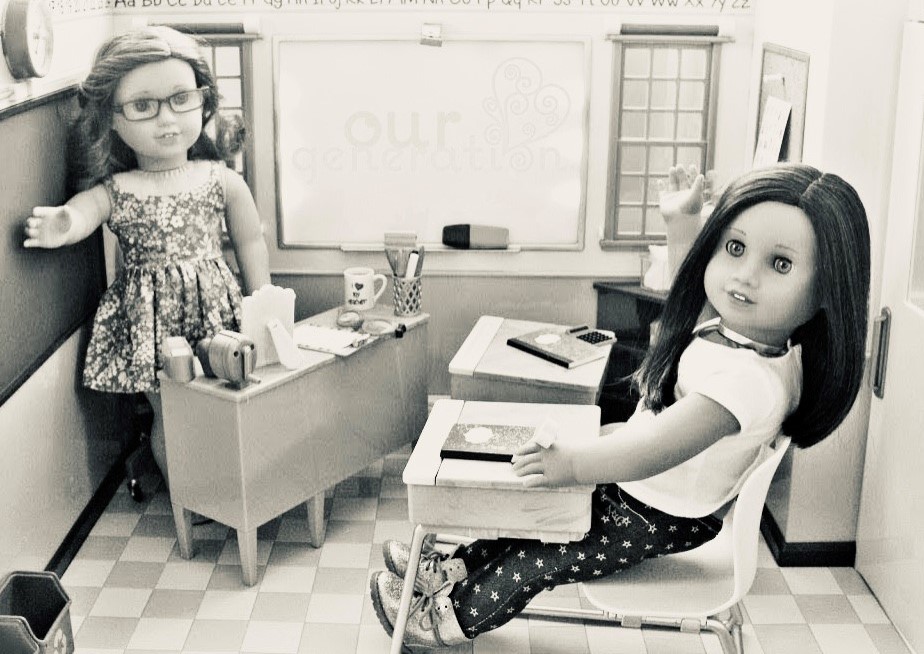 Picture shows a black and white photo of a toy classroom with dolls as teacher and student, the teacher doll stands behind a desk while the student doll sits at a desk and faces away from the teacher while raising her hand
