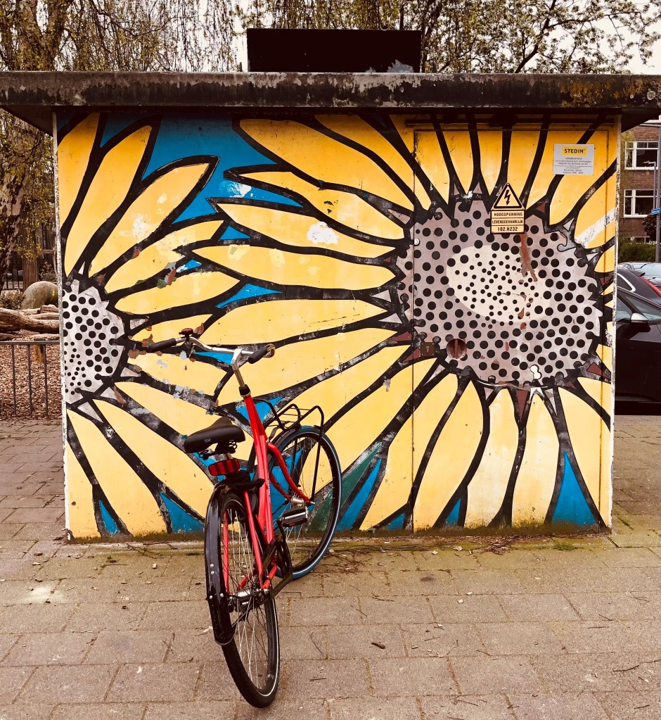 Picture shows a colour photo of a red bicycle standing on a pavement in front of an electricity substation decorated with graffiti of two large yellow sunflowers