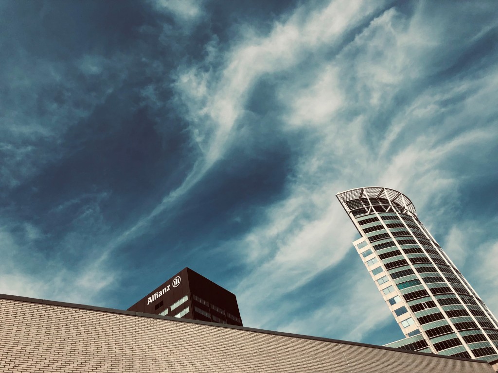 Picture shows a colour photograph of an urban skyline, in which two high rise office buildings point up into the cloudy sky from behind a windowless brick wall.