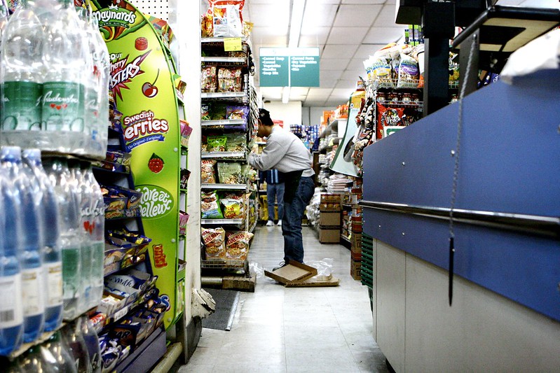 Picture shows a colour photo taken in a supermarket where a shop worker bends to place goods on the shelves stacked high with packets and bottles, while on the floor discarded packaging lays. A pen hangs from a chain on the checkout desk. The photo is entitled 'superMarket' and is courtesy of kris krüg on Flickr (CC BY-SA 2.0).