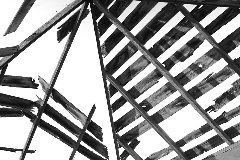 Picture is a striking black and white photo of the remains of a timber roof. The photographer is standing beneath the roof looking up. The structure of the roof is in poor condition, there are no tiles or other covering remaining, and parts of the roof timbers have fallen away, leaving a geometric pattern of right angles against a grey white sky.